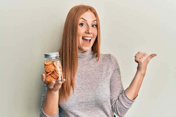 Young irish woman holding jar with salty biscuits pointing thumb up to the side smiling happy with open mouth  - Fotoğraf, Görsel