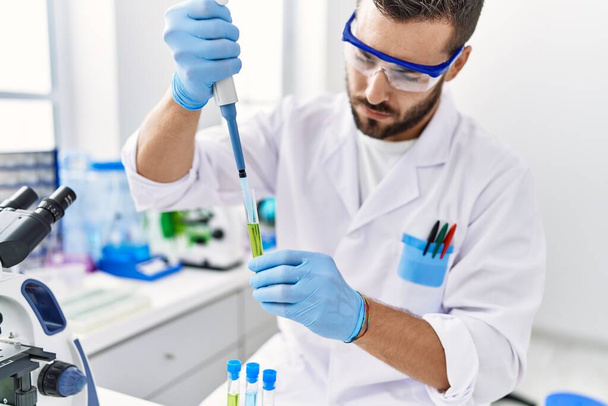 Young hispanic man wearing scientist uniform using pipette and test tube at laboratory - Photo, Image