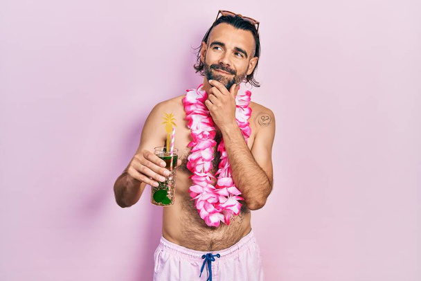 Joven hombre hispano con traje de baño y lei hawaiano bebiendo cóctel tropical con la mano en la barbilla pensando en la pregunta, expresión pensativa. Cara sonriente y pensativa. concepto de duda.  - Foto, imagen