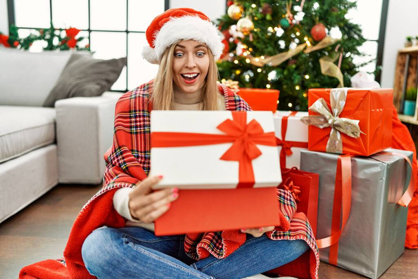jong blond vrouw unboxing gift zitten door christmas boom thuis - Foto, afbeelding