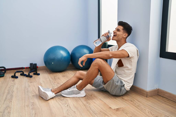 Young hispanic man drinking water sitting on floor at sport center - Zdjęcie, obraz