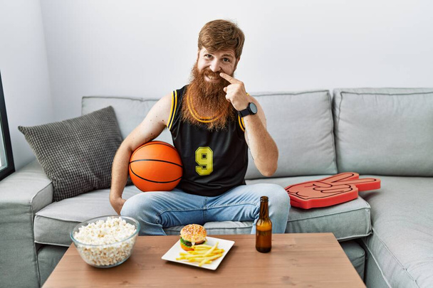 Hombre caucásico con barba larga sosteniendo pelota de baloncesto animando juego de tv señalando con la mano el dedo a la cara y la nariz, sonriendo alegre. concepto de belleza  - Foto, imagen