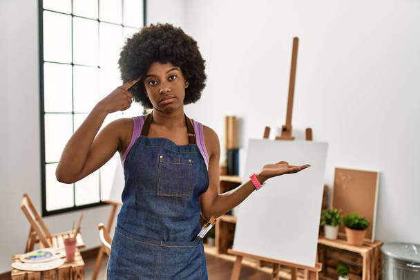 Jovem afro-americana com cabelo afro no estúdio de arte confuso e irritado com a palma aberta mostrando espaço de cópia e apontando o dedo para a testa. Pense nisso..  - Foto, Imagem