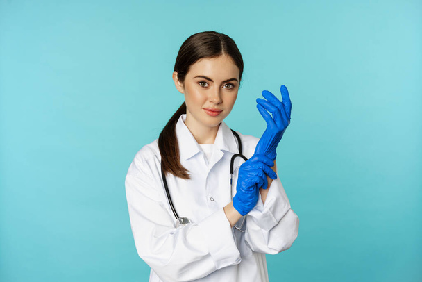 Young doctor, professional physician woman, put on gloves, smiling at camera, ready for medical checkup, standing in uniform over blue background - Photo, Image