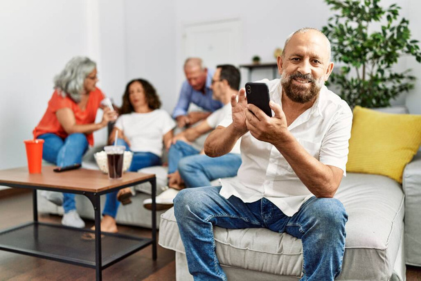 Ομάδα μεσήλικων φίλων που κάθονται στον καναπέ και μιλάνε. Ο άνθρωπος χαμογελά χαρούμενος χρησιμοποιώντας smartphone στο σπίτι. - Φωτογραφία, εικόνα