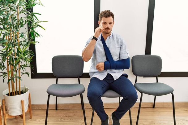 Handsome young man sitting at doctor waiting room with arm injury mouth and lips shut as zip with fingers. secret and silent, taboo talking  - Foto, Imagem