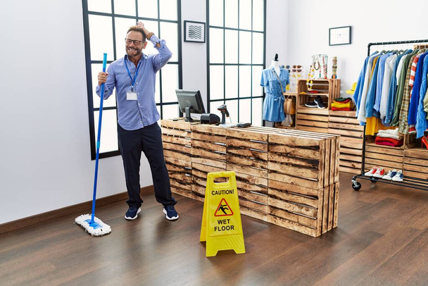 Middelbare leeftijd man werkt bij retail boetiek schoonmaken van de vloer gestresst en gefrustreerd met de hand op het hoofd, verrast en boos gezicht  - Foto, afbeelding