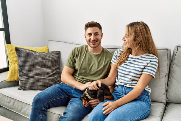 Νεαρό ζευγάρι ισπανόφωνων χαμογελά χαρούμενος κάθεται στον καναπέ με το σκυλί στο σπίτι. - Φωτογραφία, εικόνα