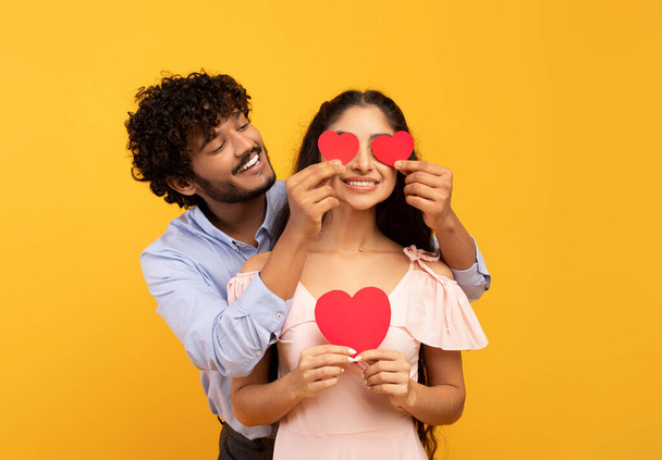 Liefdesverdriet. Liefdevolle indische echtgenoot bedekken vrouwen ogen met papier harten, poseren over gele studio achtergrond - Foto, afbeelding