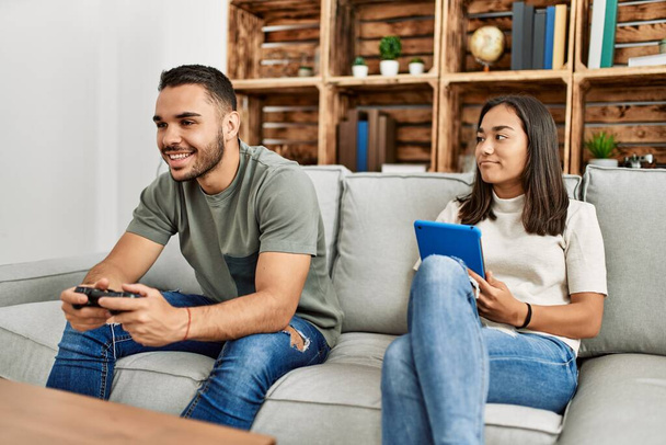 Νεαρό λατινικό ζευγάρι παίζει βιντεοπαιχνίδι και χρησιμοποιεί touchpad στο σπίτι. - Φωτογραφία, εικόνα