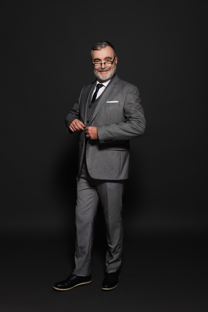 full length view of happy senior man in suit looking at camera on dark grey - Photo, image