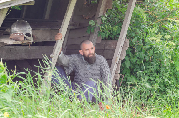 Viking warrior rest near  wooden building after the battle. Metal chain mail and helmet. Green bushes. - Foto, Bild