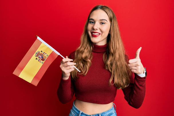 Mladá blondýnka drží španělskou vlajku s veselým a pozitivním úsměvem, palec nahoru dělá vynikající a schvalovací znamení  - Fotografie, Obrázek