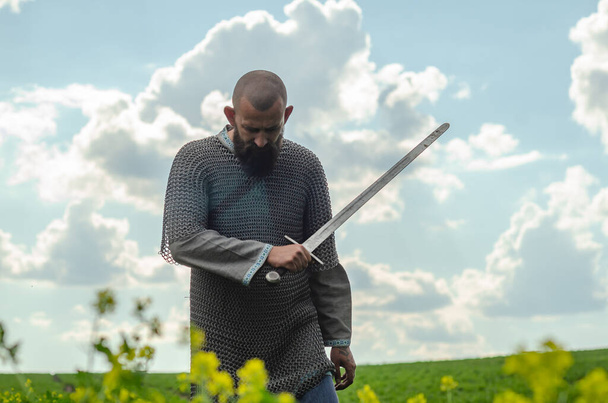 Kopasz szakállas férfi fémláncos levélben, vászoningen áll a mező közepén, karddal a kezében. Harcias tekintet. Homályos háttér. - Fotó, kép