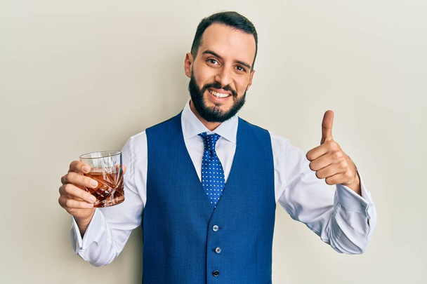 Молодой человек с бородой пьет виски выстрел улыбаясь счастливым и позитивным, большой палец вверх делает отлично и знак одобрения  - Фото, изображение
