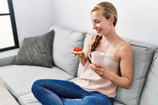 Young caucasian woman having breakfast sitting on sofa at home - Photo, Image