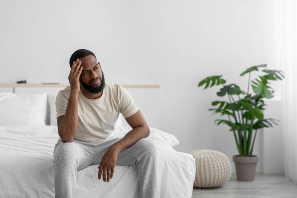 Desperate young african american guy with beard suffers from loneliness, depression in bedroom interior - 写真・画像