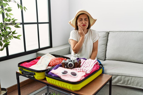 Beautiful middle age hispanic woman packing summer clothes in suitcase touching mouth with hand with painful expression because of toothache or dental illness on teeth. dentist  - Valokuva, kuva