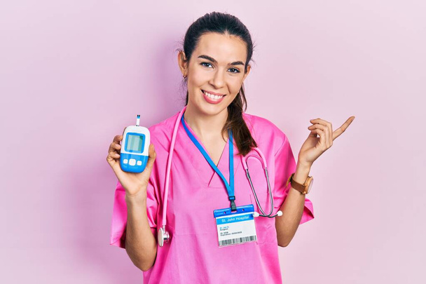Young brunette woman wearing doctor uniform holding glucometer smiling happy pointing with hand and finger to the side  - Zdjęcie, obraz