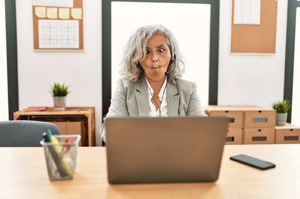 Middle age businesswoman sitting on desk working using laptop at office making fish face with lips, crazy and comical gesture. funny expression.  - Photo, image