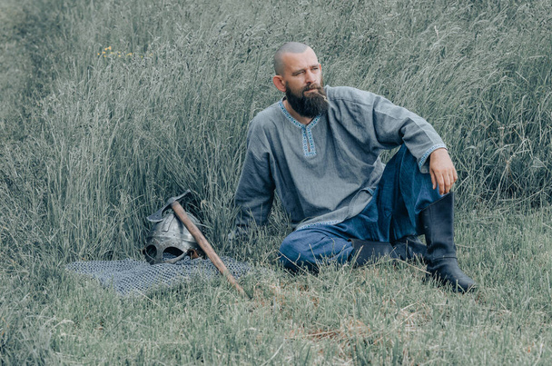 Мужественный человек с бородой, покоящийся на траве в поле. Страшный взгляд. Рядом с ним топор и шлем воина. - Фото, изображение