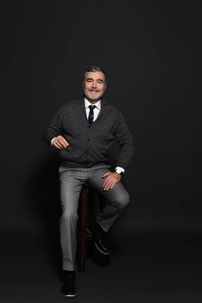 full length view of senior man in stylish clothes sitting on high stool and smiling at camera on grey - Fotografie, Obrázek