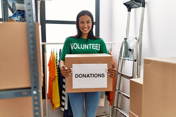 Önkéntes egyenruhát viselő fiatal latin nő adománygyűjtő dobozt tart a jótékonysági központban. - Fotó, kép