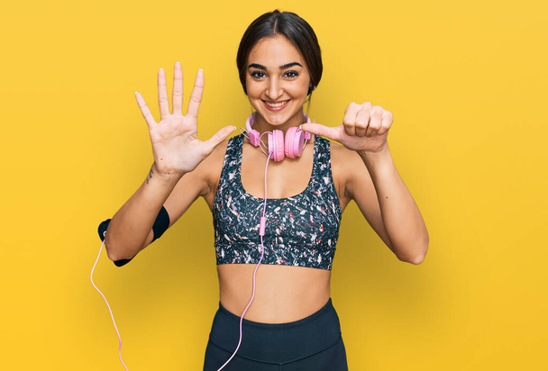 Beautiful brunette woman wearing gym clothes and using headphones showing and pointing up with fingers number six while smiling confident and happy.  - Fotoğraf, Görsel