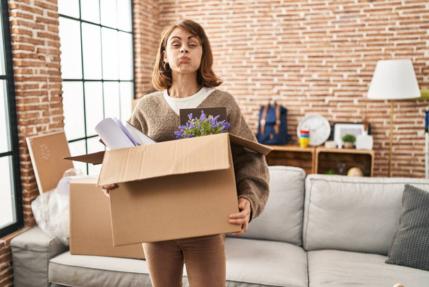 Young beautiful woman holding box moving to a new home puffing cheeks with funny face. mouth inflated with air, catching air.  - Foto, afbeelding