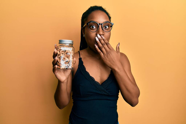 African american woman with braided hair holding walnuts covering mouth with hand, shocked and afraid for mistake. surprised expression  - Фото, зображення