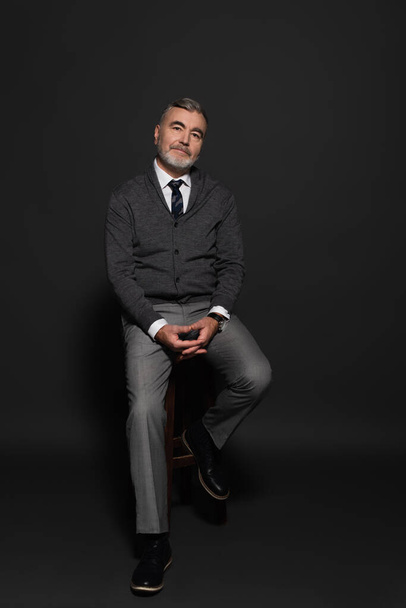 full length view of smiling grey-haired man in stylish clothes sitting on high stool on dark grey - Φωτογραφία, εικόνα
