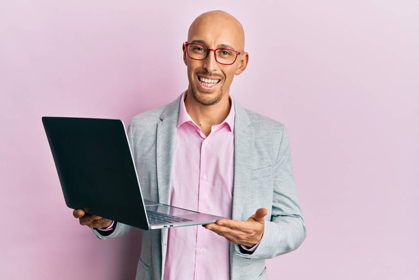Bald man with beard holding laptop looking positive and happy standing and smiling with a confident smile showing teeth  - Fotó, kép