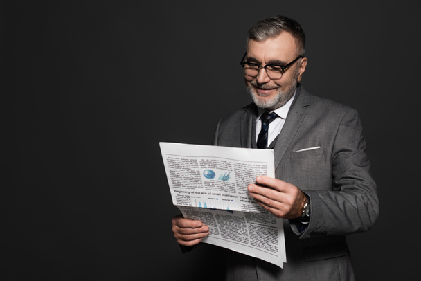 happy senior man in suit and eyeglasses reading newspaper isolated on dark grey - Foto, Imagen