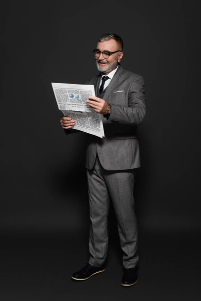 full length of senior man in suit and eyeglasses reading newspaper on dark grey - Zdjęcie, obraz
