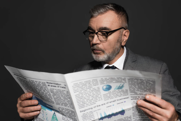 bearded senior businessman in eyeglasses reading blurred newspaper isolated on dark grey - Fotó, kép