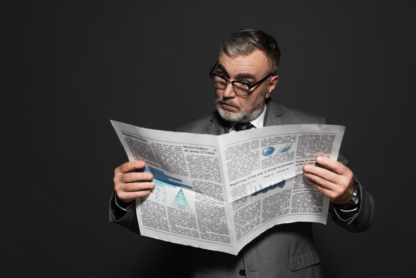 surprised businessman in eyeglasses reading newspaper isolated on dark grey - Fotó, kép