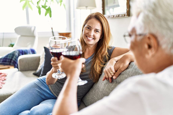 Μεσήλικες ισπανόφωνοι ζευγάρι χαμογελάνε ευτυχισμένοι πίνοντας κρασί καθισμένοι στον καναπέ στο σπίτι. - Φωτογραφία, εικόνα