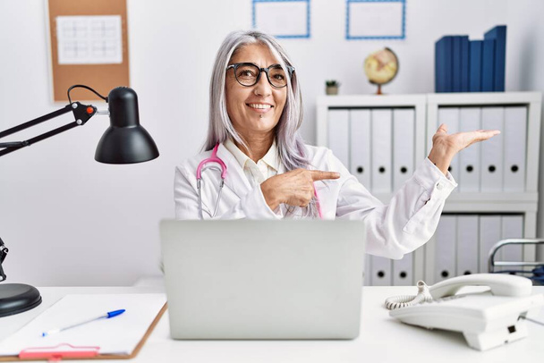 Mujer de pelo gris de mediana edad vistiendo uniforme médico trabajando con computadora portátil sorprendida y sonriendo a la cámara mientras se presenta con la mano y señalando con el dedo.  - Foto, imagen