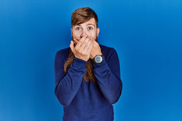 Redhead man with long beard wearing casual blue sweater over blue background shocked covering mouth with hands for mistake. secret concept.  - Foto, immagini