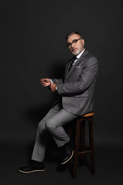 full length view of senior man in suit and eyeglasses sitting on high stool and looking at camera on dark grey - Foto, Bild