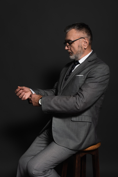 senior man adjusting sleeve of blazer while sitting on stool on dark grey - Fotó, kép