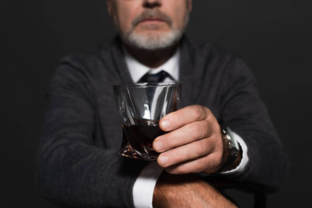 cropped view of blurred man in grey jumper holding glass of whiskey isolated on black - Foto, afbeelding