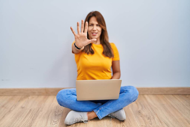 Mujer hispana usando laptop sentada en el suelo en casa haciendo stop gesture con palmas de las manos, expresión de enojo y frustración  - Foto, imagen