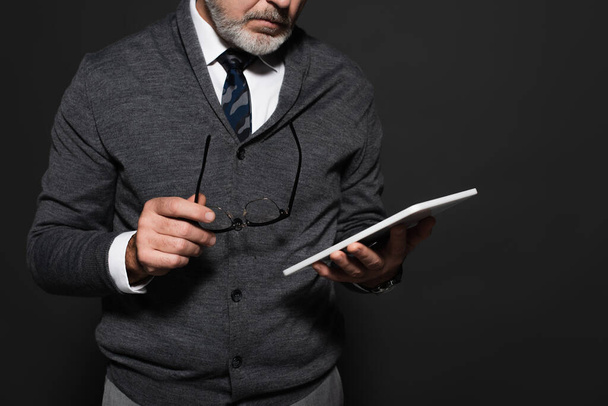 cropped view of senior man in stylish clothes holding eyeglasses and digital tablet isolated on dark grey - 写真・画像