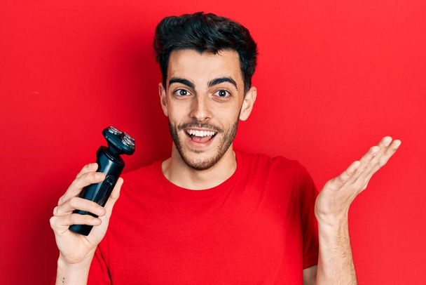 Joven hombre hispano sosteniendo máquina de afeitar eléctrica celebrando el logro con sonrisa feliz y expresión ganadora con la mano levantada  - Foto, Imagen