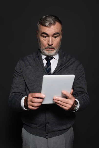 surprised senior man in jumper and tie looking at digital tablet isolated on dark grey - Zdjęcie, obraz