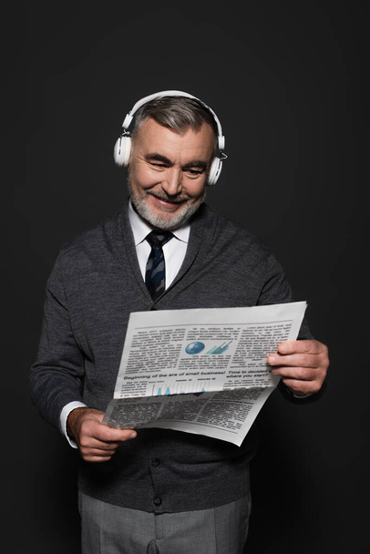 happy senor man in jumper, tie and headphones reading newspaper on dark grey - Valokuva, kuva