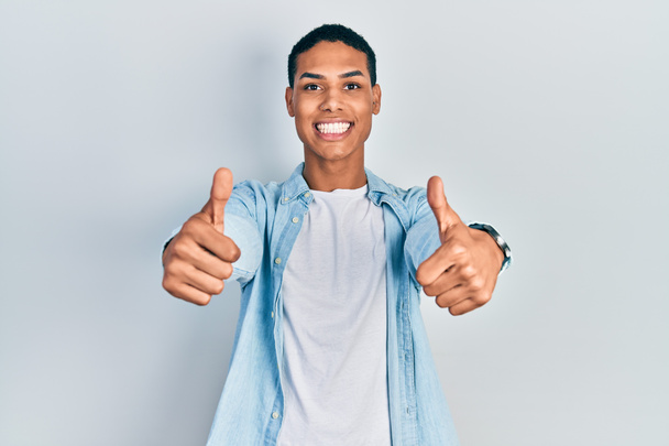 Genç Afro-Amerikalı adam günlük giysiler giyip olumlu jestler yapıyor. Elleriyle gülümsüyor ve başarı için seviniyor. kazanan jest.  - Fotoğraf, Görsel