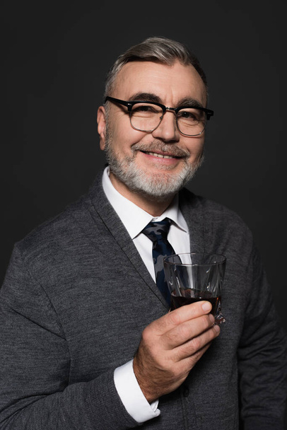positive senior man in eyeglasses holding glass of whiskey while looking at camera isolated on dark grey - Foto, imagen