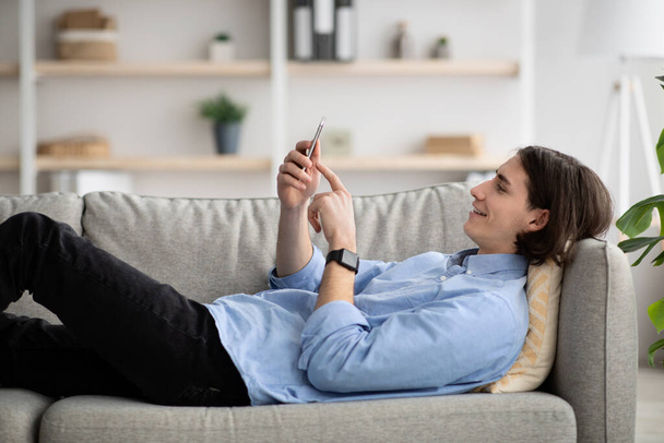 Человек наслаждается свободное время в одиночестве лежа на диване и смотреть видео на мобильном телефоне, расслабляясь дома, используя технику - Фото, изображение